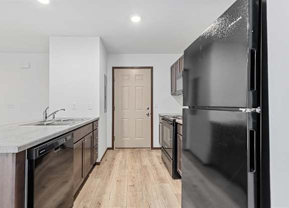 A black refrigerator is in the foreground of a kitchen with wooden floors and white walls.