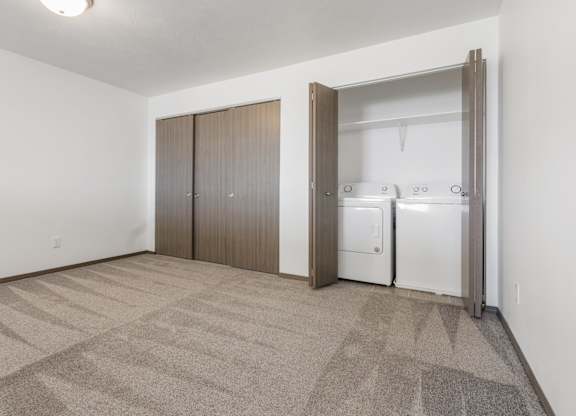 A room with a carpeted floor and a white wall with a fridge and cupboards in it.