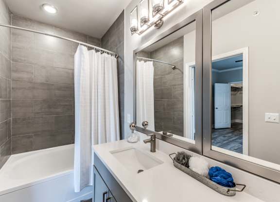 A modern bathroom with a white tub, sink, and grey tiled walls at Rosemary Apartments, Conroe, TX, 77384