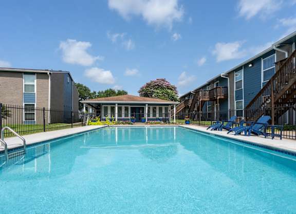 A swimming pool in front of a building with a patio and chairs.