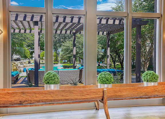 A wooden bench with three green plants on it in front of a window with a view of a pool and trees.