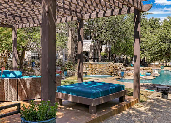 A wooden pergola over a sunbed with a pool in the background.