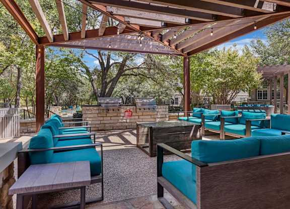 A wooden pergola with blue cushioned seating is surrounded by trees.