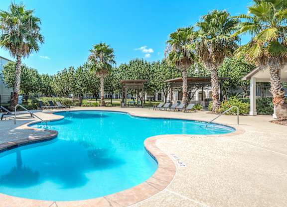 A large swimming pool surrounded by palm trees and a white building at Rosemary Apartments, Conroe, Texas