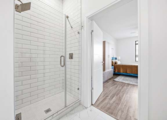 A white tiled bathroom with a walk-in shower and a wooden floor leading to a bedroom.
