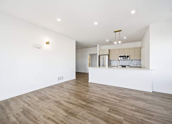 A room with wooden floors and a kitchen area in the background.