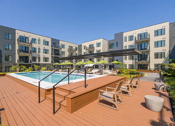 A wooden deck with a pool and lounge chairs in front of apartment buildings.