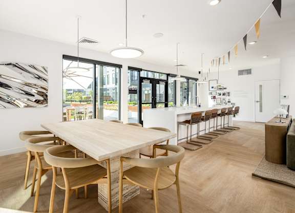 A long wooden table is surrounded by chairs in a white room with a large window.