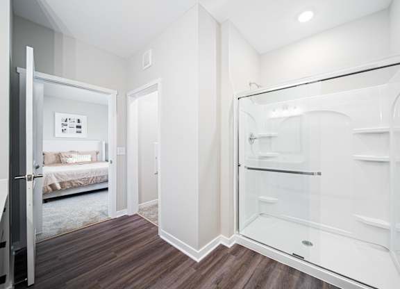 A white bathroom with a walk-in shower and a bedroom visible through the door.