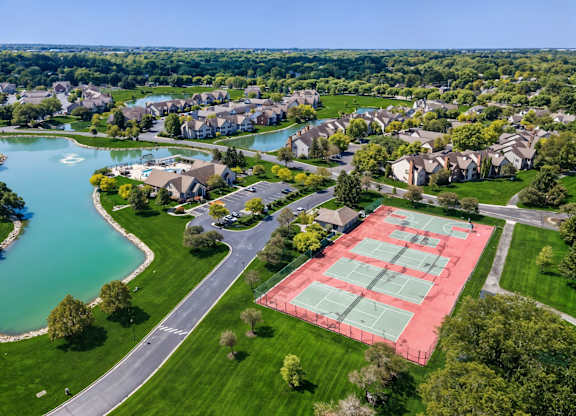 A tennis court is located in a grassy area near a lake and houses.