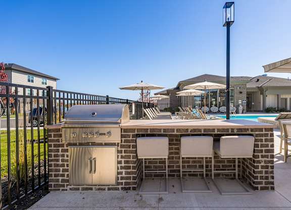 A brick barbecue grill with two chairs and an umbrella in front of a pool.