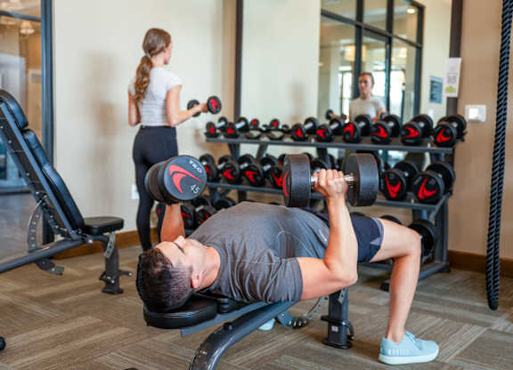 A man is working out in a gym with a woman in the background.