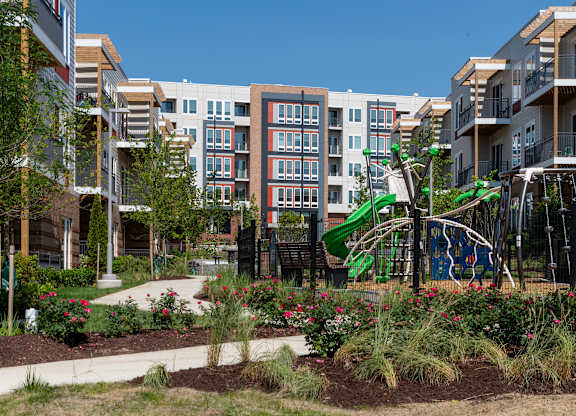 A playground with a green slide and a white swing set is surrounded by a garden with red flowers and green shrubs.