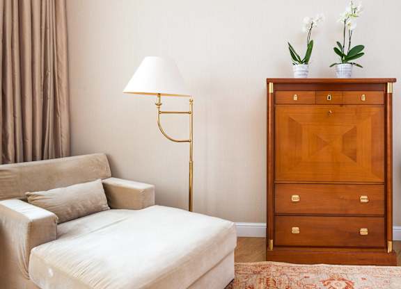 A white sofa sits in front of a wooden chest of drawers with a lamp and two potted plants beside it.