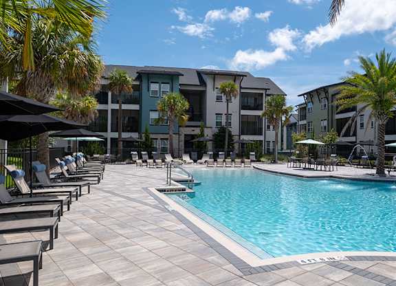 A swimming pool surrounded by lounge chairs and umbrellas in front of a row of apartment buildings.