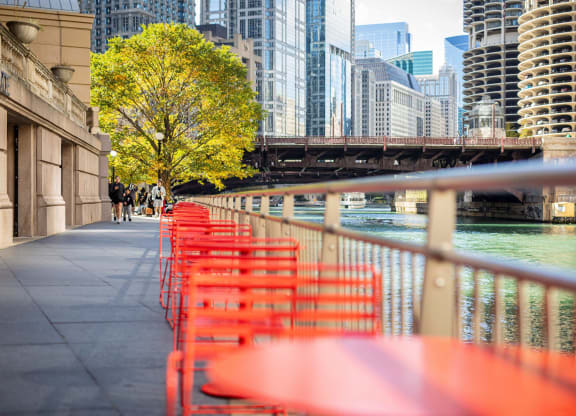a row of red tables and chairs next to the river in a city at Columbus Plaza, Chicago, IL, 60601