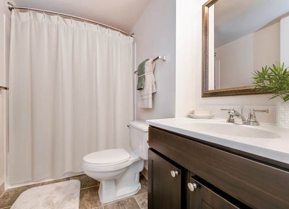 A white toilet sits in a bathroom with a white shower curtain and a mirror above a sink.