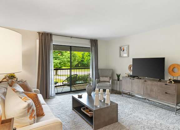 A living room with a white couch, a coffee table, a television, and a sliding glass door at The Waverly on the Lake - Belleville, MI Apartment , Belleville