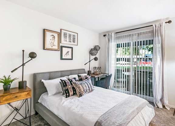 A bedroom with a bed, a nightstand, and a window with a view of a balcony at The Waverly on the Lake - Belleville, MI Apartment , Michigan, 95050