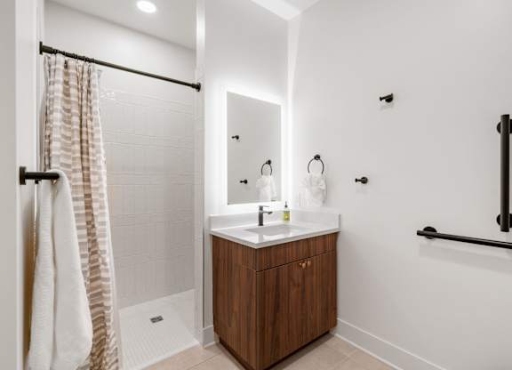 A bathroom with a vanity, backlit mirror, toilet, and walk-in shower in the Guest Suite, a reservable community amenity at Lilia in Lilydale, MN 55118