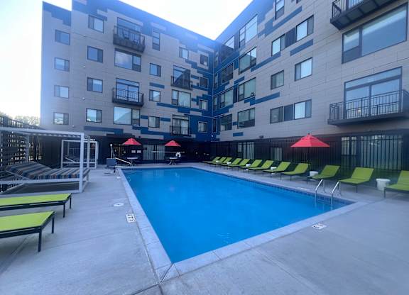 A large swimming pool surrounded by lounge chairs and umbrellas in front of a multi-story apartment building.
