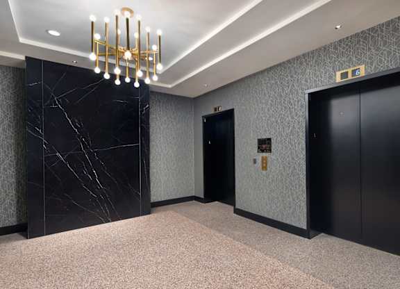A hallway with a chandelier, marble wall, and two elevators.