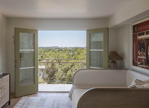 A bedroom with a bed, dresser, and a balcony with a view of the trees at The PARKER on E Mulberry Apartments, San Antonio