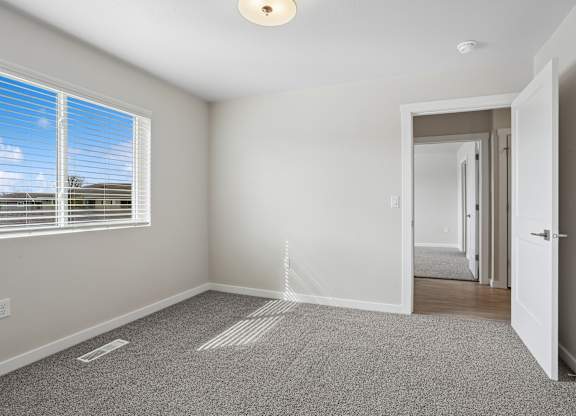 A room with a carpeted floor, a window with blinds, and a door leading to another room.
