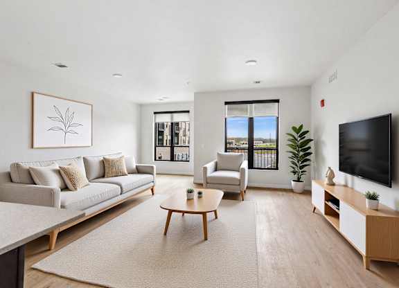 A living room with a grey couch, a wooden coffee table, a flat screen TV, and a large window with a view of the outside.