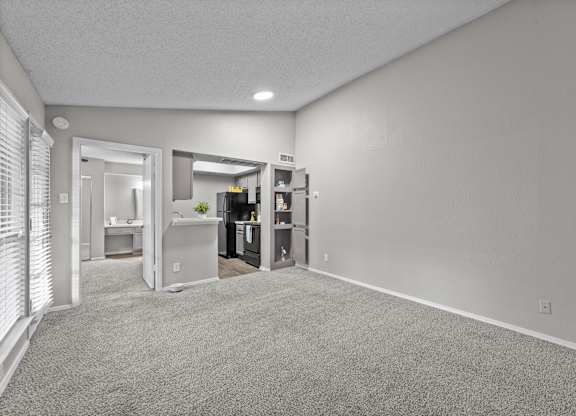 A spacious room with a carpeted floor and a kitchen area visible through the open door.