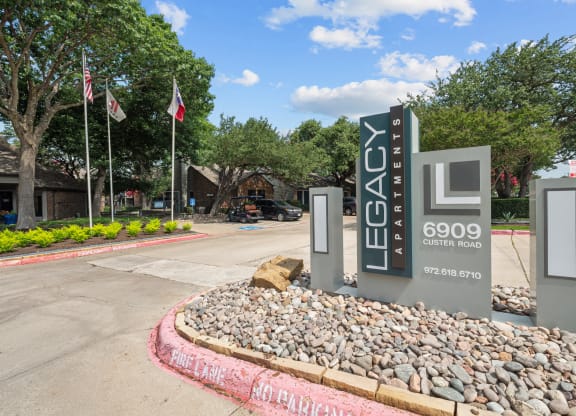 a sign that says Legacy Apartments at the entrance to a parking lot with flags in the background
