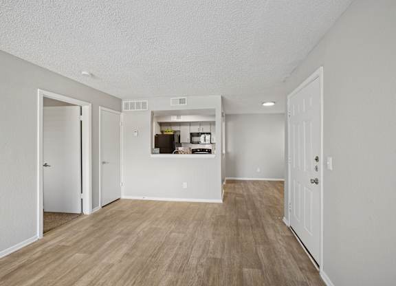 A room with white walls and wooden flooring with a kitchen visible through a doorway.