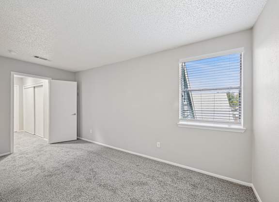 A room with a carpeted floor, a window with blinds, and a door leading to another room.