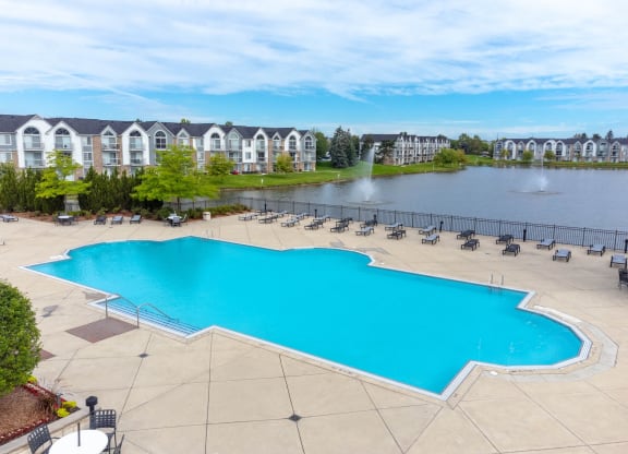 a large swimming pool with chaise lounge chairs next to a lake with apartment buildings in theat The Harbours Apartments, Clinton Twp, MI