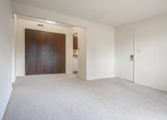 an empty living room with white carpet and a wooden door at Bavarian Village Apartments, Indianapolis, IN