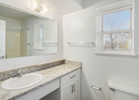 A bathroom with a sink, mirror, toilet, and a window with blinds at Black Sand Apartment Homes in Lincoln, NE
