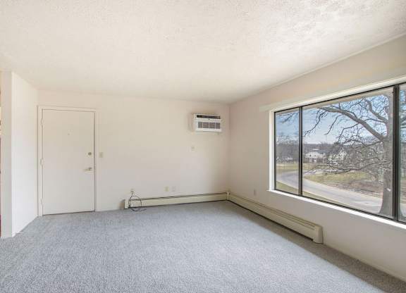 A living room with a grey carpet and a window showing a tree at Concord Place Apartments, Kalamazoo, MI, 49009