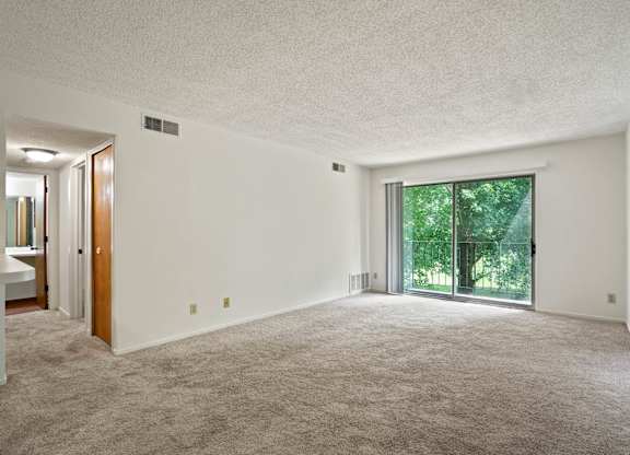 A large, empty room with a carpeted floor and a sliding glass door leading to a green outdoor area at Beacon Hill and Great Oaks Apartments, Rockford, Illinois