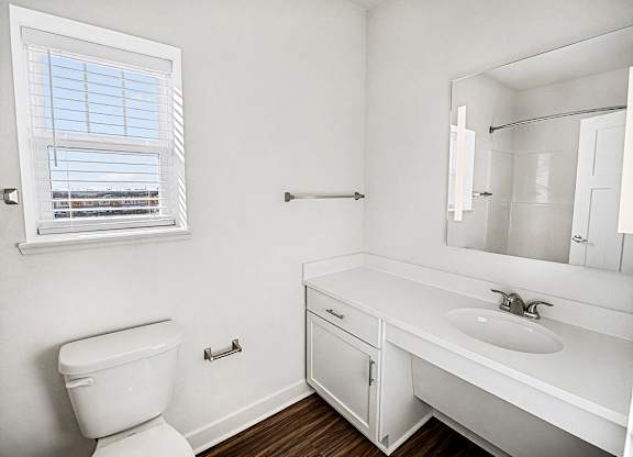 A white bathroom with a toilet, sink, mirror and a window at The Grove Apartment Homes in Lincoln, NE
