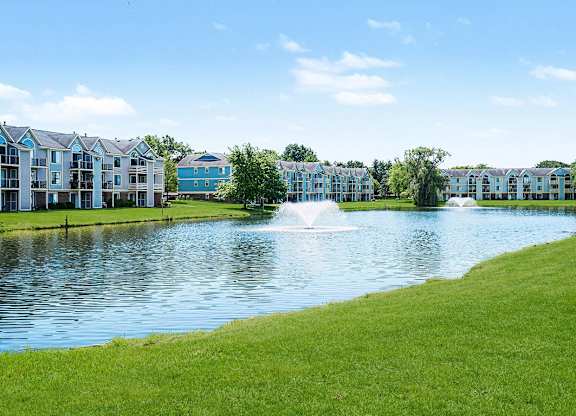 A serene pond with a fountain in the middle of a grassy area with buildings in the background at Indian Lakes Apartments, Mishawaka, Indiana