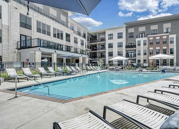 A swimming pool surrounded by sun loungers and umbrellas in front of apartment buildings at Luxe 360 Apartment Complex in Midlothian Virginia