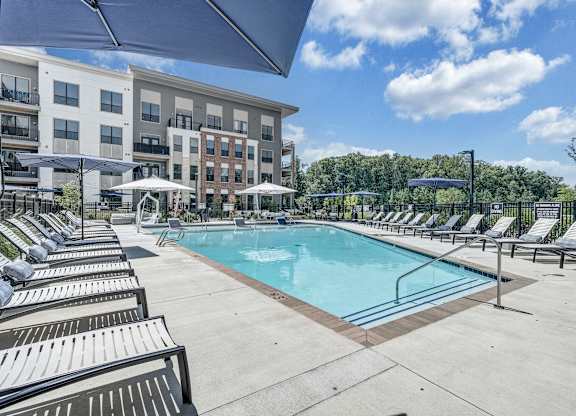 A swimming pool surrounded by sun loungers and umbrellas in front of apartment buildings at Luxe 360 Apartment Complex in Midlothian Virginia