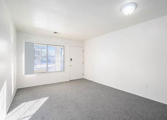 A living room with a carpeted floor, a window, and a door at Newport Village Apartments, Portage, MI