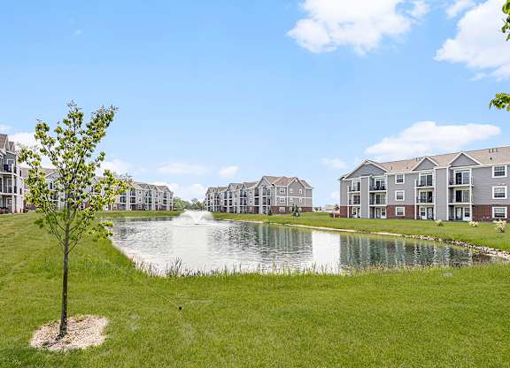 A tree stands in front of a pond with green grass behind apartment buildings. at Tracy Creek Apartments, Perrysburg