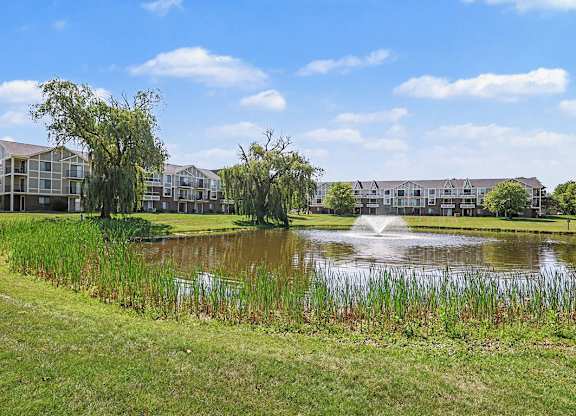A pond in the middle of a grassy area with a building in the background at Trappers Cove Apartments, Lansing, MI, 48910