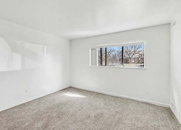 A garden style living room with a carpeted floor and a window showing a view of trees outside at Trappers Cove Apartments, Lansing