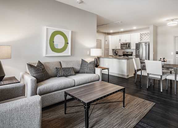A modern living room with a grey couch, a coffee table, and a dining area in the background.