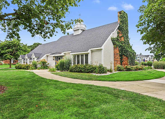 A community building with a grey roof and a chimney is surrounded by green grass and trees at Walnut Trail Apartments, Michigan