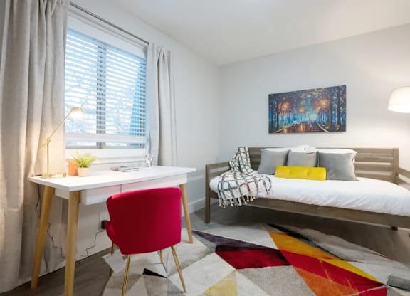 A room with a red chair, a white desk, and a white bed with a colorful rug.