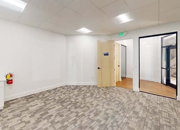 A room with a carpeted floor, a white wall, a wooden door, and a white cabinet with a red object on top.